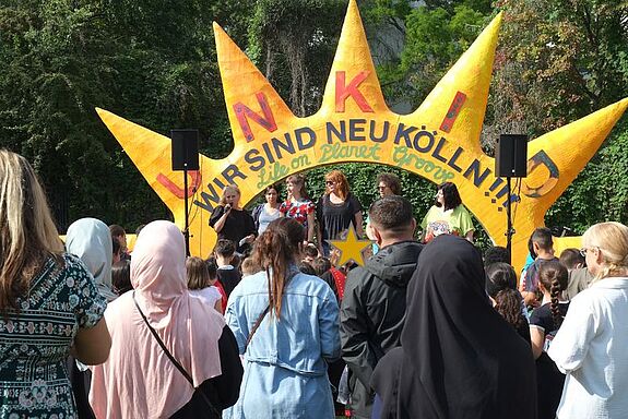 Bühne mit großer gelber Dekoration in Form einer Sonne und dem Schriftzug „Wir sind Neukölln“, davor zahlreiche Kinder und Erwachsene.
