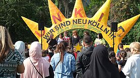 Bühne mit großer gelber Dekoration in Form einer Sonne und dem Schriftzug „Wir sind Neukölln“, davor zahlreiche Kinder und Erwachsene.