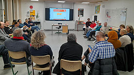 Eine Gruppe von Menschen sitzt im Kreis und unterhält sich. Am Kopf der Runde hängt ein Monitor, auf dem eine Präsentation läuft. 
