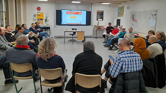 Eine Gruppe von Menschen sitzt im Kreis und unterhält sich. Am Kopf der Runde hängt ein Monitor, auf dem eine Präsentation läuft. 