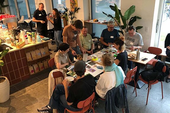 Mehrere Personen sitzen und stehen in einem Café um einen Tisch und arbeiten gemeinsam mit Karten und Fotos an Ideen für den Quartiersrat.