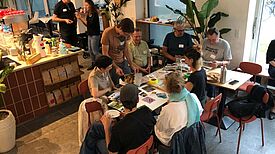 Mehrere Personen sitzen und stehen in einem Café um einen Tisch und arbeiten gemeinsam mit Karten und Fotos an Ideen für den Quartiersrat.