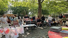 Menschen stehen an einer mobilen Siebdruckwerkstatt auf einem Platz unter Bäumen neben einem Wäscheständer, auf dem bedruckte Blätter und T-Shirts trocknen, und Tischen mit Arbeitsmaterialien.