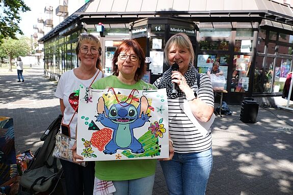 Drei Frauen stehen vor einem Gebäude, in den Händen halten Sie ; eine bunte Geschenktüte und ein Mikrofon.