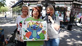 Drei Frauen stehen vor einem Gebäude, in den Händen halten Sie ; eine bunte Geschenktüte und ein Mikrofon.