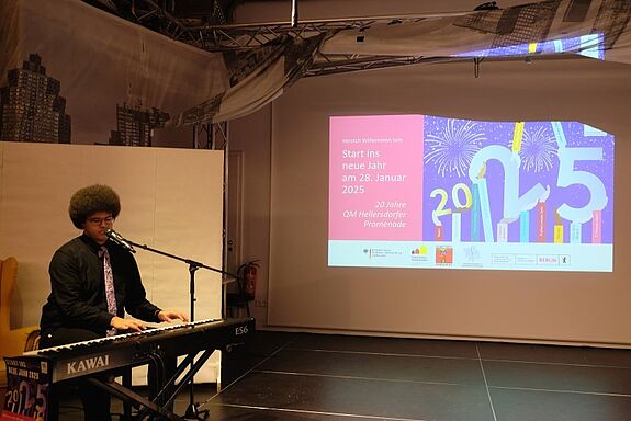 Der Musiker Ilya Ananyin spielt Klavier vor einer Leinwand mit der Aufschrift „Start ins neue Jahr – 20 Jahre QM Hellersdorfer Promenade“.