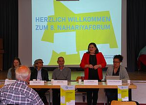 Fünf Vertreterinnen aus Politik, Verwaltung und Quartiersmanagement vor einer Präsentation, die das Publikum zum achten Nahariyaforum begrüßen.