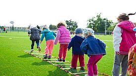 Kinder spielen gemeinsam auf einem Sportplatz und üben eine Koordinationsübung auf einer Balancierleiter.