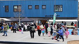 Menschen besuchen ein Nachbarschaftsfest vor einem blauen Gebäude, Kinder spielen, Erwachsene stehen an Ständen und kommen ins Gespräch.