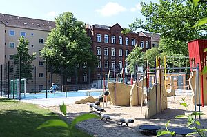 Spielplatz mit Kletter- und Balanciergeräten aus Holz und Seilen, daneben ein eingezäuntes Spielfeld mit blauem Boden. Im Hintergrund ein Wohnhaus und ein rotes Backsteingebäude sowie Bäume.