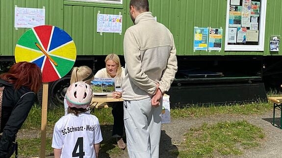Kinder und Eltern stehen vor dem Bauwagen an einem Glücksrad-Stand von Schwarz-Weiß Spandau im Falkenhagener Feld.