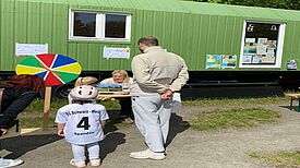 Kinder und Eltern stehen vor dem Bauwagen an einem Glücksrad-Stand von Schwarz-Weiß Spandau im Falkenhagener Feld.
