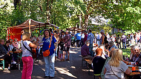Menschen sitzen und stehen an langen Bänken und Marktständen unter Bäumen bei der Kaffeetafel 2025 in der Gropiusstadt Nord.