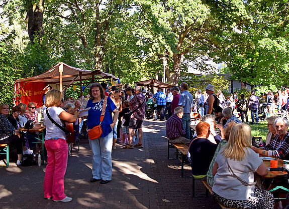 Menschen sitzen und stehen an langen Bänken und Marktständen unter Bäumen bei der Kaffeetafel 2025 in der Gropiusstadt Nord.