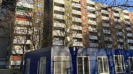 Auf dem Bild sieht man ein Hochhaus, vor dem das temporäre Stadtteilzentrum in einem blauen Containerbau untergebracht sein wird.  