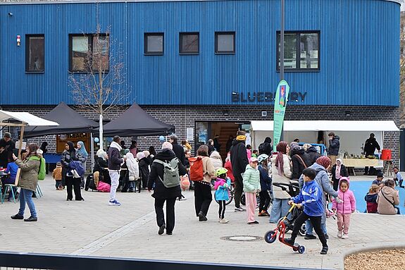 Menschen besuchen ein Nachbarschaftsfest vor einem blauen Gebäude, Kinder spielen, Erwachsene stehen an Ständen und kommen ins Gespräch.