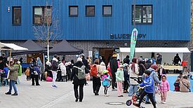 Menschen besuchen ein Nachbarschaftsfest vor einem blauen Gebäude, Kinder spielen, Erwachsene stehen an Ständen und kommen ins Gespräch.