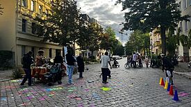 Kinder und Erwachsene spielen zwischen bunten Kreidezeichnungen und Pylonen auf einer Pflasterstraße.