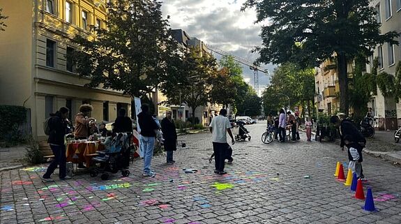 Kinder und Erwachsene spielen zwischen bunten Kreidezeichnungen und Pylonen auf einer Pflasterstraße.