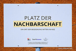 Weißes Schild vor ockerfarbenem Hintergrund mit der Aufschrift „Platz der Nachbarschaft. Ein Ort der Begegnung im Kiez“. Darunter Logos vom Quartiersmanagement Falkenhagener Feld Ost, Berlinovo und casa e.V. und die Erläuterung, dass der Platzname nicht offiziell, sondern von der Nachbarschaft als Zeichens des Miteinanders vergeben wurde. 