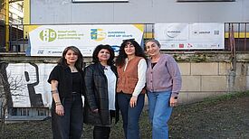 Vier Frauen posieren vor einem Gebäude und lächeln in die Kamera.