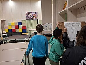 Vier Kinder von hinten sichtbar schreiben Namen auf ein Papier an einer Pappwand. Auf dem Papier steht „Open-Stage-Liste, Singen, Tanzen, Karaoke. Im Hintergrund Regale, eine Bühne, bunte Sitzwürfel, ein Schild „Moabiter Kinderhof“ an der Wand.