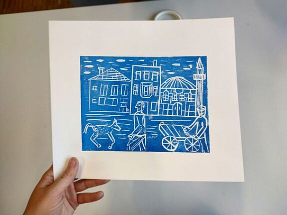 Eine Hand hält einen Kunstdruck auf dem Häuser sowie Menschen zu sehen sind. 