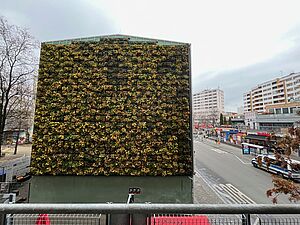 Draufsicht auf eine bepflanzte Gebäudefassade im urbanen, stark versiegelten Umfeld. Sichtbar neben dem begrünten Gebäude sind ein Baum, eine Straße mit Busspur und Wohnhochhäuser aus den 1970er Jahren.