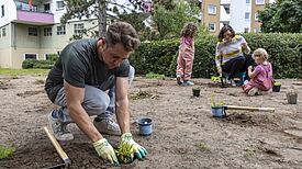 Ein Mann pflanzt auf einer offenen Fläche mit Gartenhandschuhen eine Staude ein; im Hintergrund sind zwei Kinder und eine Frau.
