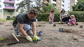 Ein Mann pflanzt auf einer offenen Fläche mit Gartenhandschuhen eine Staude ein; im Hintergrund sind zwei Kinder und eine Frau.