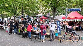 Menschen sitzen und stehen an langen Tischen auf der Hellersdorfer Promenade, bunte Wimpel hängen zwischen den Bäumen.
