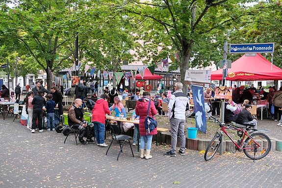 Menschen sitzen und stehen an langen Tischen auf der Hellersdorfer Promenade, bunte Wimpel hängen zwischen den Bäumen.