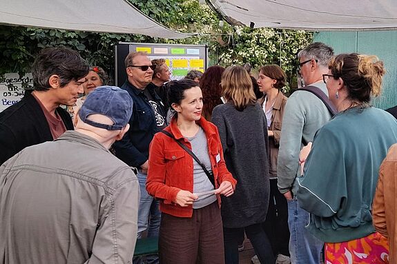 Mehrere Menschen stehen im Gespräch beisammen bei einer öffentlichen Veranstaltung im Freien. Im Hintergrund ist ein Infoboard mit bunten Zetteln zu sehen. Eine Frau in roter Jacke spricht mit Teilnehmenden.