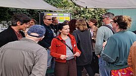 Mehrere Menschen stehen im Gespräch beisammen bei einer öffentlichen Veranstaltung im Freien. Im Hintergrund ist ein Infoboard mit bunten Zetteln zu sehen. Eine Frau in roter Jacke spricht mit Teilnehmenden.