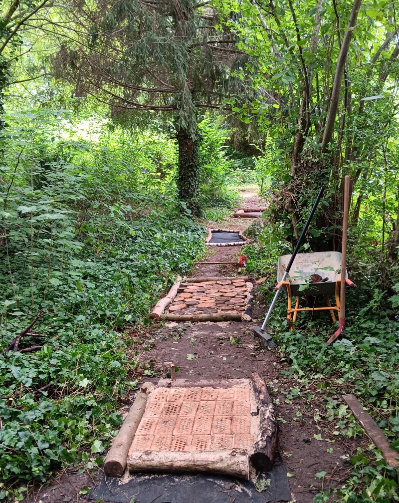 Vier Barfußpfadflächen befüllt mir verschiedenen Naturmaterialien führen durch einen dichten, grünen Park; daneben stehen Schubkarre, Rechen und Schaufel.