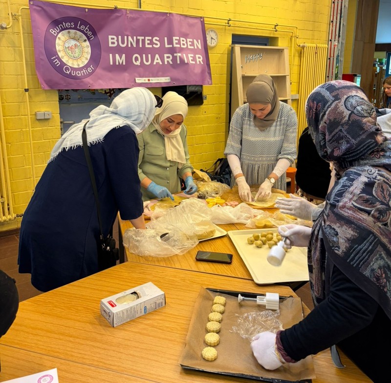 Mehrere Frauen bereiten an einem Tisch in einem Raum an Tischen kleine Gebäckstücke zu. Im Hintergrund hängt ein Banner, auf dem „Buntes Leben im Quartier“ steht.