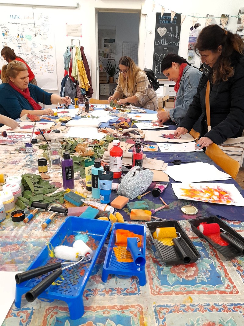 Mehrere Personen sitzen an einem großen Tisch und bedrucken Stoffbeutel mit buntem Herbstlaub. Auf dem Tisch liegen Farben, Pinsel, Walzen und bemalte Blätter.