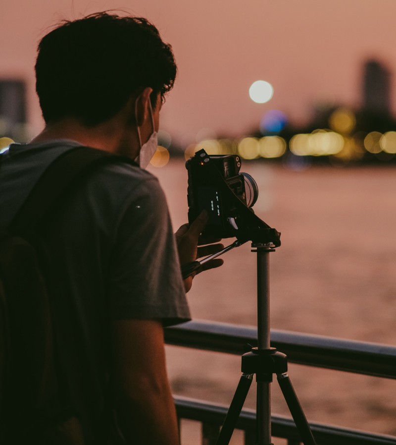Eine Person fotografiert mit einer Kamera auf einem Stativ den Abendhimmel über einer unscharfen Stadtansicht.