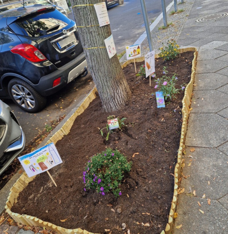 Eine frisch bepflanzte, mit Holzkante eingefasste Baumscheibe am Straßenrand ist mit bunten Blumen und selbstgemalten Hinweisschildern von Kindern zum Thema Umweltschutz und Sauberkeit gestaltet.