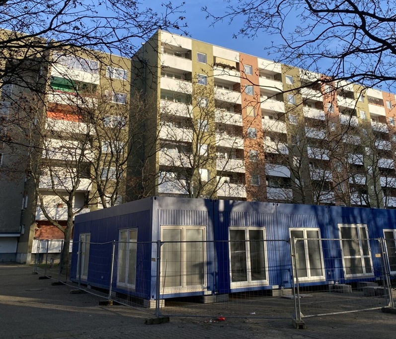 Auf dem Bild sieht man ein Hochhaus, vor dem das temporäre Stadtteilzentrum in einem blauen Containerbau untergebracht sein wird.  