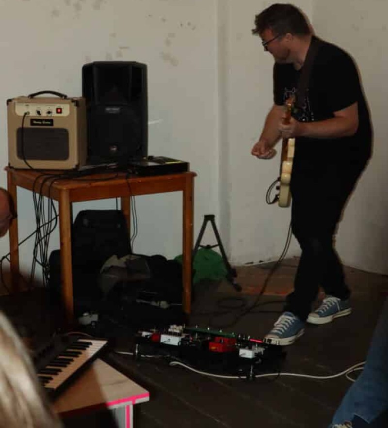 Ein Mann steht mit einer elektronischen Gitarre an einem Verstärker und Lautsprecher; Effektgeräte und ein kleines Keyboard liegen auf dem Boden.