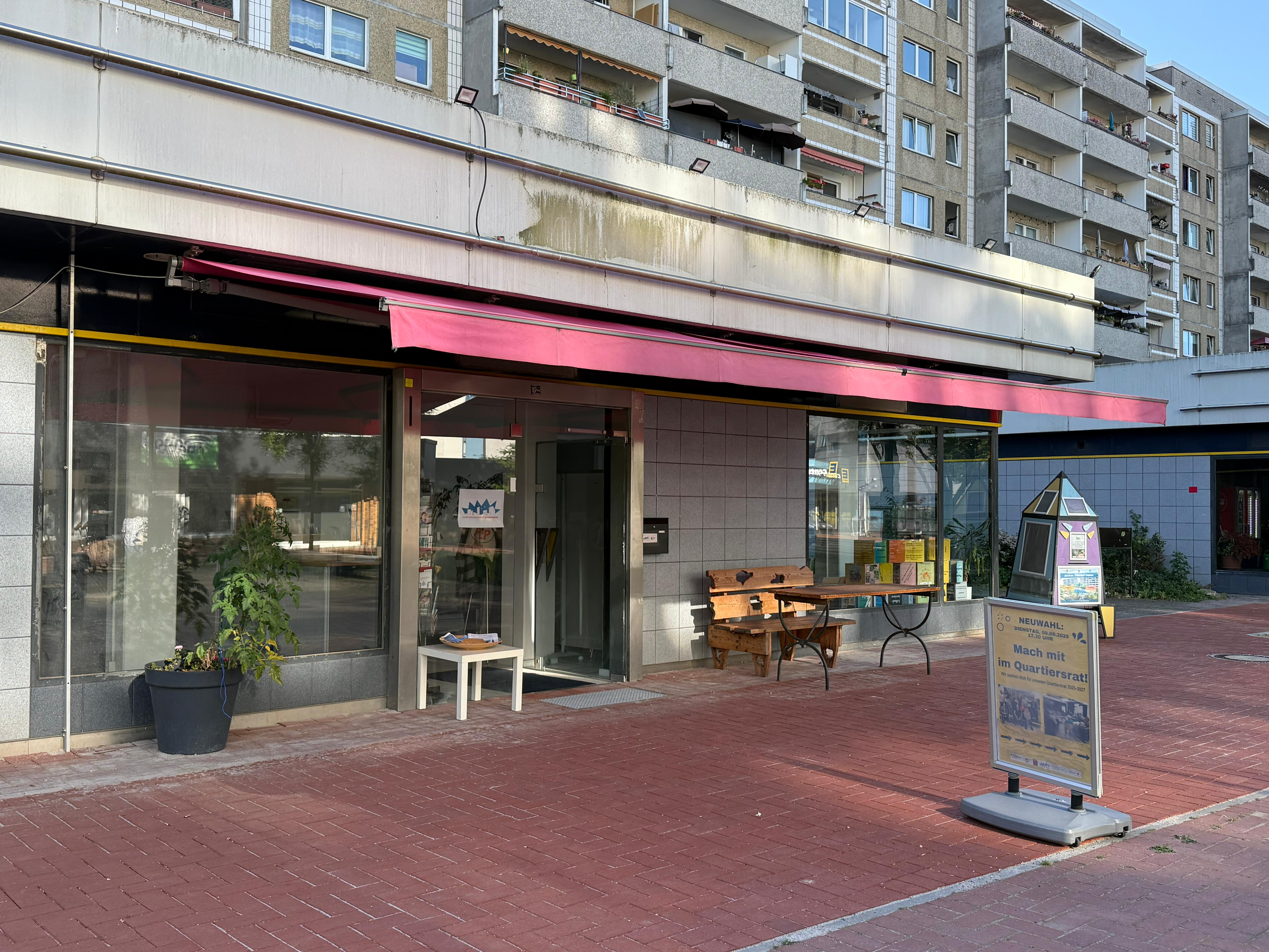 Außenansicht des QM-Büros mit Bank und Informationsaufsteller davor.