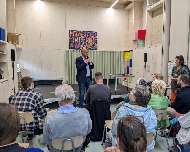 Staatssekretär Stephan Machulik steht vor eine Gruppe von Menschen und hält Rede. 