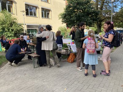 Menschen sitzen und stehen an Tischen im Freien, Kinder spielen, während Pizza am mobilen Kiezofen gebacken wird.