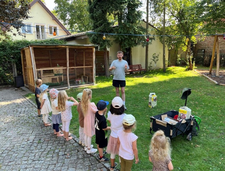 Kinder stehen in einer Reihe im Kita-Garten und hören einem Mann zu, der eine Pflanzaktion erklärt.