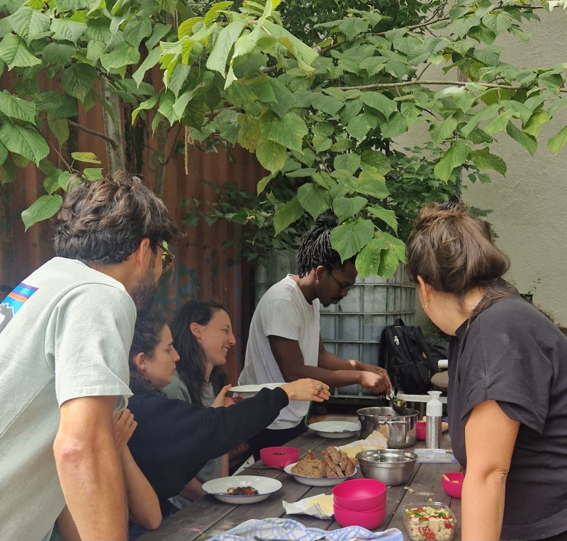 Fünf Personen sitzen oder stehen um einen Tisch im Freien unter einem Baum. 