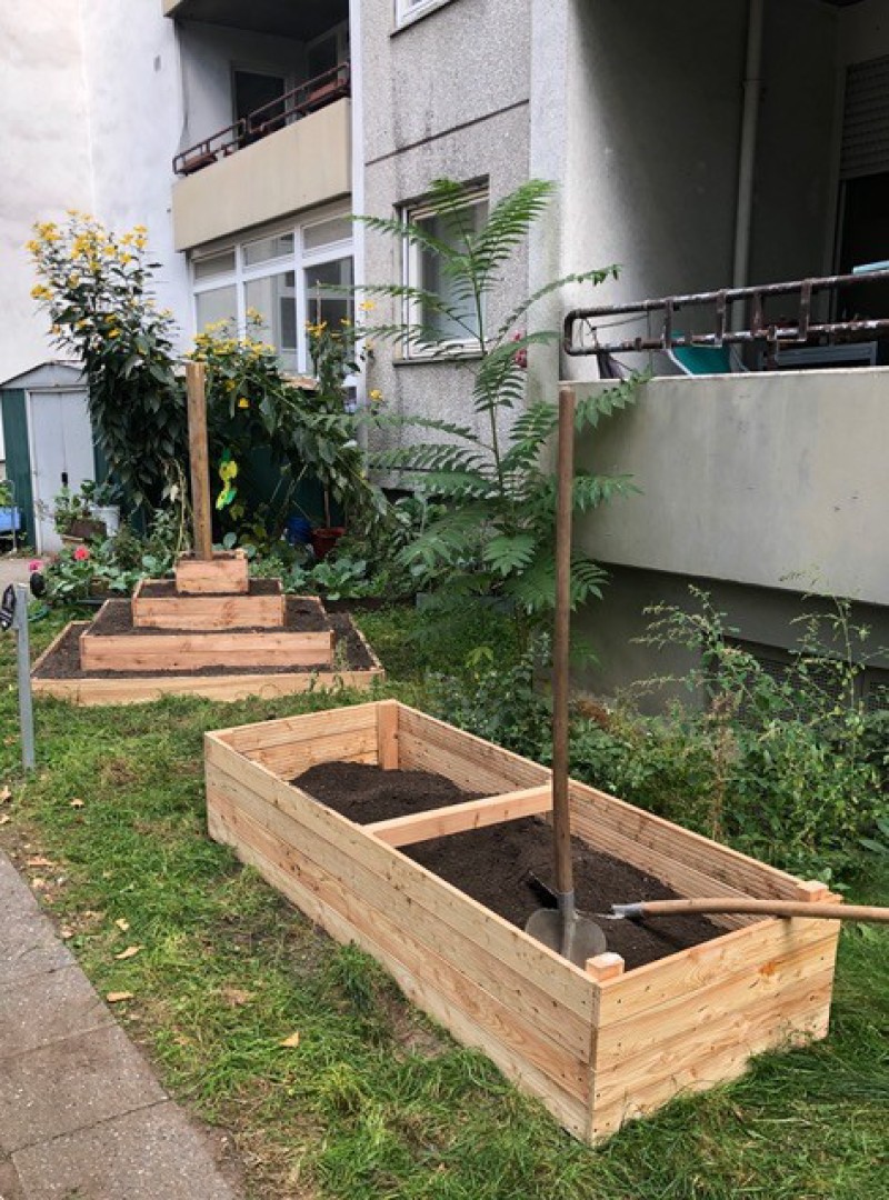 Zwei Schaufeln liegen auf einem mit Erde befüllten Hochbeet vor einem Wohnhaus. Daneben steht ein Pyramidenbeet.