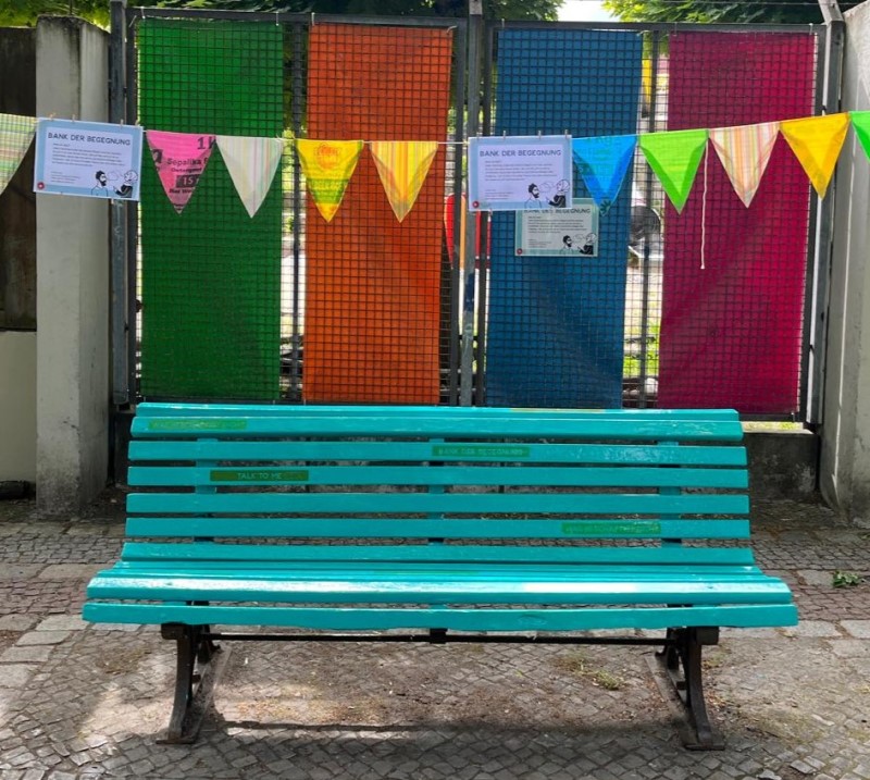 Türkisfarbene Bank auf dem Falkplatz vor bunter Wimpelgirlanden, Teil des Projekts „Bank der Begegnung“ in der Rollbergsiedlung.